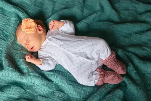 Малышка с вязаной повязкой с цветком на голове и шерстяных носках спит на пледе